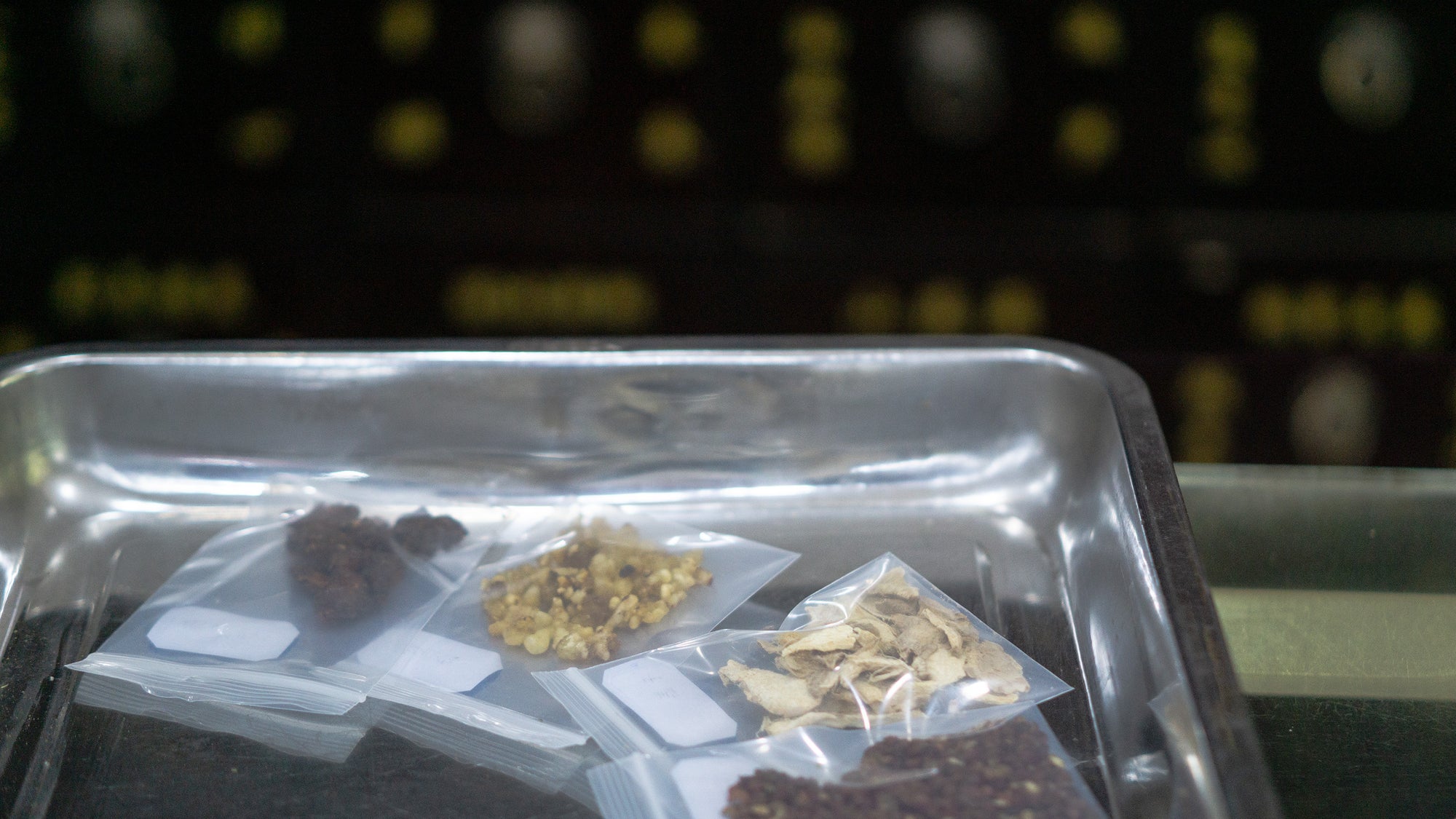 Small portions of dried botanical ingredients sealed in clear sample bags, arranged in a metal tray during preparation.