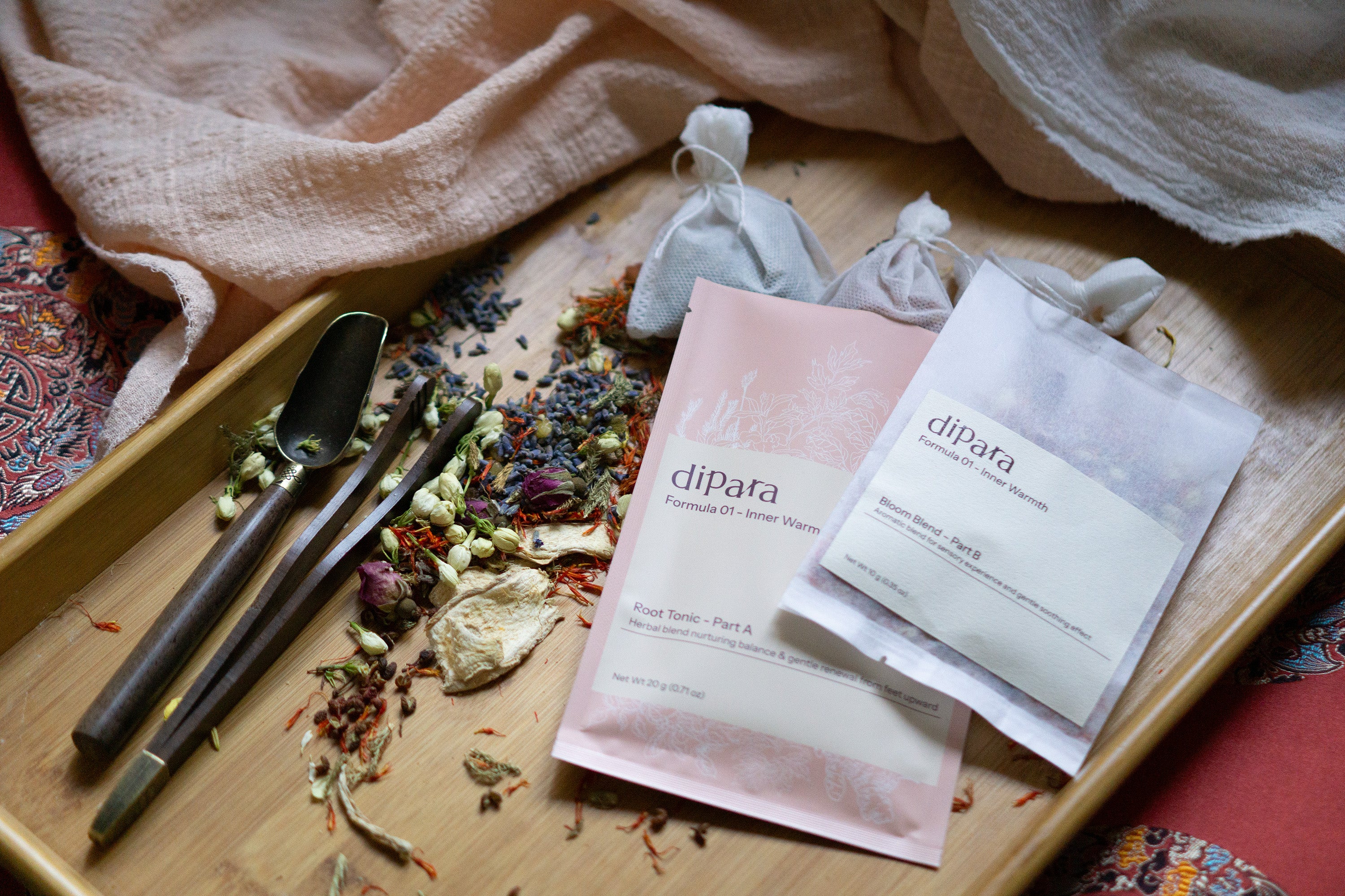 Dipara Formula 01 Inner Warmth two-part herbal foot soak sachets displayed with dried botanicals and tools on a wooden tray.